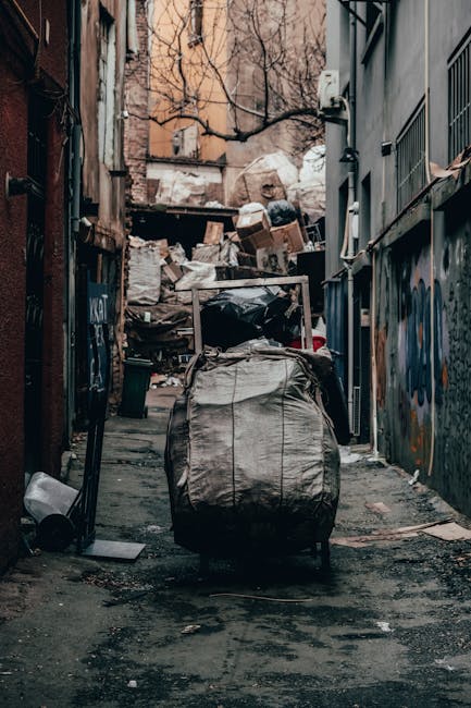 A narrow alleyway between brick and concrete buildings during daytime, with a large, gray, fabric-covered trolley or cart positioned centrally on the pavement, used for moving bulky waste or furniture collection. In the background, piles of assorted cardboard boxes, black plastic bags, and other waste materials are stacked against a fence, indicating ongoing waste removal or a home relocation staging area. The alley features graffiti on the metal doors and walls, with some exposed pipework and wiring visible on the building facades. The scene captures the process of loading or transporting large items, possibly related to clearance or renovation, with natural lighting illuminating the space. Man with Van Rayners Lane offers services related to removals, packing, and furniture transport, as suggested by the context of the image and the environment for home or building clearance.