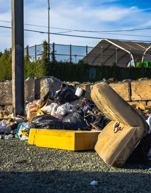 A collection of bulky waste including a large, worn out beige sofa and various black and plastic bags filled with household rubbish, placed on the pavement outside a property. The sofa, with visible fabric covering, is partially disassembled and leaning against a pile of miscellaneous discarded items. An open yellow plastic box is positioned nearby among the debris, which also contains cardboard boxes and packaging materials. In the background, a stone wall, a fence, and taut overhead power lines are visible, with a clear blue sky overhead. The scene indicates a waste removal process typically associated with house clearance or moving logistics, relevant to services provided by Man with Van Rayners Lane for bulky waste disposal and home relocation efforts.