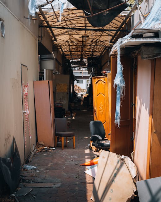 A narrow alleyway between brick and concrete buildings during daytime, with a large, gray, fabric-covered trolley or cart positioned centrally on the pavement, used for moving bulky waste or furniture collection. In the background, piles of assorted cardboard boxes, black plastic bags, and other waste materials are stacked against a fence, indicating ongoing waste removal or a home relocation staging area. The alley features graffiti on the metal doors and walls, with some exposed pipework and wiring visible on the building facades. The scene captures the process of loading or transporting large items, possibly related to clearance or renovation, with natural lighting illuminating the space. Man with Van Rayners Lane offers services related to removals, packing, and furniture transport, as suggested by the context of the image and the environment for home or building clearance.