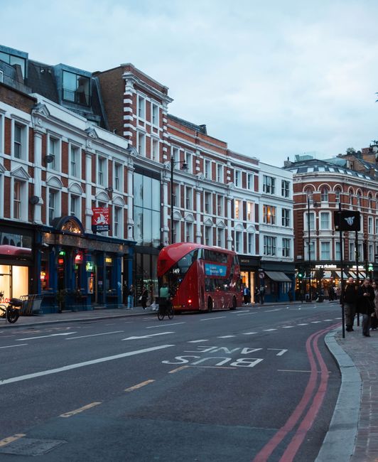 The image shows a busy outdoor shopping street on a sunny day, with numerous pedestrians walking along the pavement and browsing storefronts. On the left side, there are market stalls displaying an ar