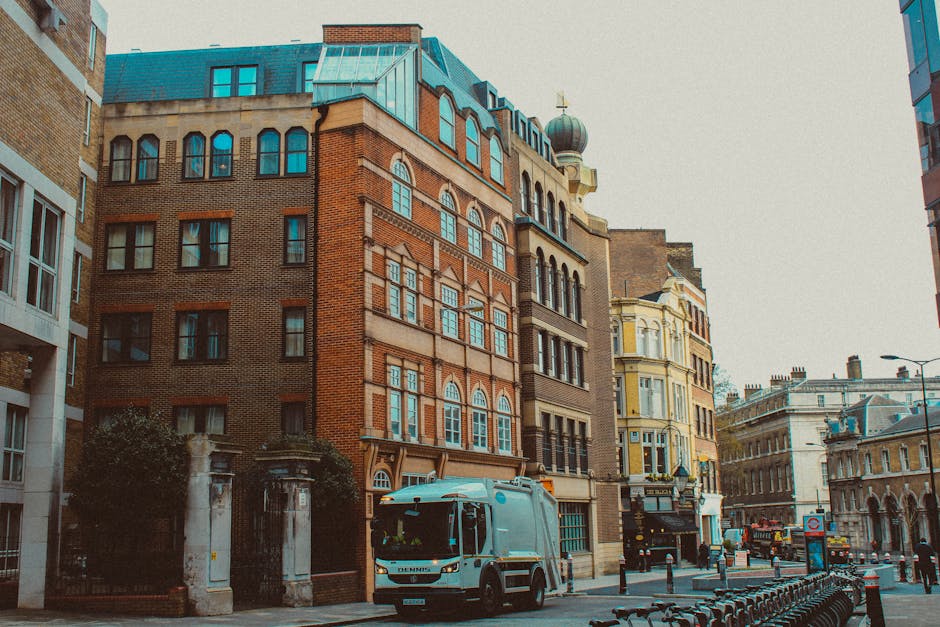 A grey van with the logo 'Demi's' is parked on the street near a row of multi-storey buildings featuring a mix of brick and ornate architectural details, typical of London. The van is positioned close to the pavement, and the rear doors are partially open as a team from Man with Van Rayners Lane is loading large, wrapped furniture and cardboard boxes onto the vehicle, indicating a home relocation or furniture transport process. The street is lined with metal barriers, and there are bicycles parked nearby. The surroundings include a lamppost, a bus stop sign, and other vehicles further down the road, with soft daylight illuminating the scene, reflecting typical urban moving logistics associated with house removals in Harrow and the careful packing and loading operations handled by professionals.