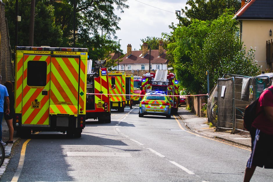 A row of emergency service vehicles, including a yellow and red London Ambulance and multiple fire trucks with flashing blue lights, are parked along a residential street in Rayners Lane. The scene is cordoned off with police tape, indicating an active emergency situation. Several personnel and bystanders are present, with some individuals standing on the pavement, observing the scene. To the right, temporary fencing made of metal mesh and fabric covers encloses part of a driveway or property boundary. The street is lined with trees, houses with red tiled roofs, and utility poles, with natural daylight illuminating the area. In the foreground, a person wearing a pink shirt and black shorts is visible on the right side, partially out of frame. The emergency vehicles are positioned close together, occupying most of the street, which appears to be temporarily closed for safety and access control. The scene reflects a coordinated emergency response, possibly linked to a building or home incident requiring urgent intervention and clearance by authorities.