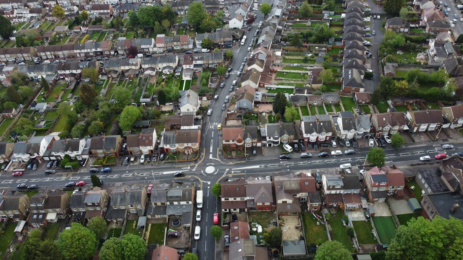 An aerial view of a residential neighbourhood showing a variety of terraced and semi-detached houses with different roof styles, located along a curved road with a roundabout at the intersection. Cars are parked along the street and in driveways, and the area is surrounded by green trees, gardens, and backyards. The scene is captured during daylight with natural lighting, highlighting the layout of the houses and the street network. This image reflects a typical urban residential area where house removals and home relocation services, such as those provided by Man with Van Rayners Lane, might be involved in logistics, packing, or furniture transport processes.