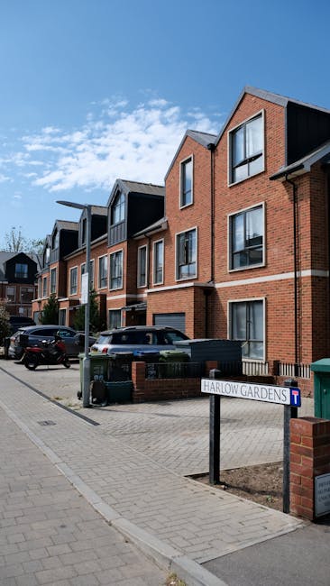 A street view in a residential area showing a modern multi-storey brick building with large windows and dark boarding on the upper floors. Several parked cars are lined up along the curb, and a black rubbish bin is placed near the building. In the foreground, there is a small sidewalk with a metal street sign reading 'Harlow Gardens' and a blue plastic bin. The sky is clear with scattered clouds, indicating a bright day. This setting depicts a typical urban neighbourhood where house removals and furniture transport might take place, with the building's entrance and nearby pavement prepared for loading or unloading activities associated with home relocation services offered by companies like Man with Van Rayners Lane.