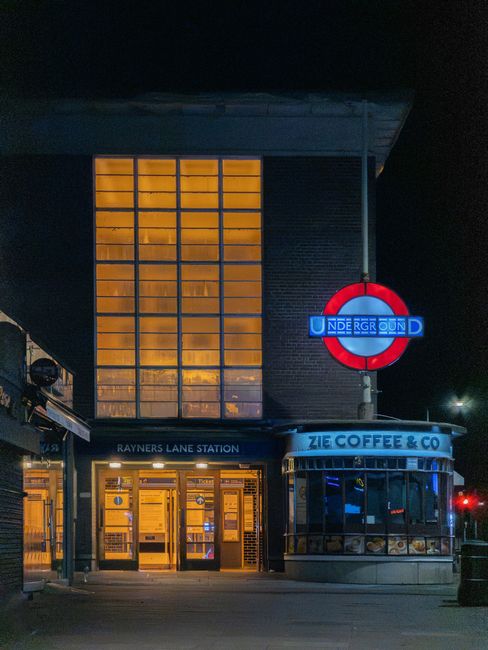 Inside a cafe, a customer with a black backpack and a puffy jacket stands at the counter, facing away from the camera. The counter displays various coffee-making equipment, including a black coffee ma