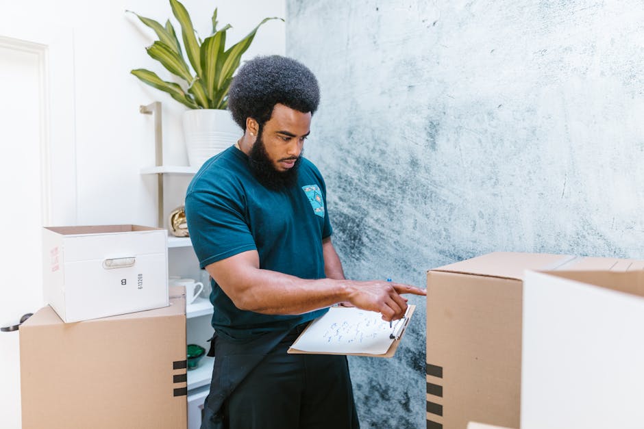 A professional mover from Man with Van Rayners Lane is inside a property during a home relocation, dressed in a navy t-shirt and black trousers, holding a clipboard and inspecting packed boxes. The bo