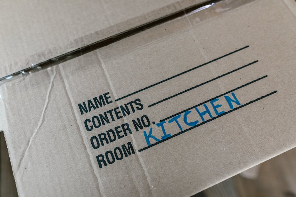A professional mover from Man with Van Rayners Lane is inside a property during a home relocation, dressed in a navy t-shirt and black trousers, holding a clipboard and inspecting packed boxes. The bo