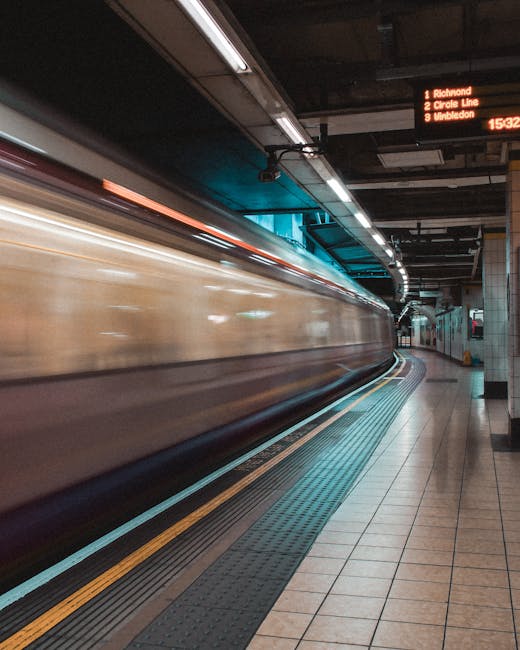 A blurred image of a moving train passing through an underground station platform with beige and black exterior, capturing motion during a home relocation process. The platform features tiled flooring with tactile paving along the edge for safety, and a digital departure board displaying train times and destinations such as Richmond, Circle Line, and Wimbledon. Overhead lighting illuminates the station, and the station walls and ceiling are lined with dark panels. This footage exemplifies the arrival or departure phase in furniture transport and packing and moving services conducted by Man with Van Rayners Lane, highlighting the importance of efficient logistics during house removals near Rayners Lane Station.