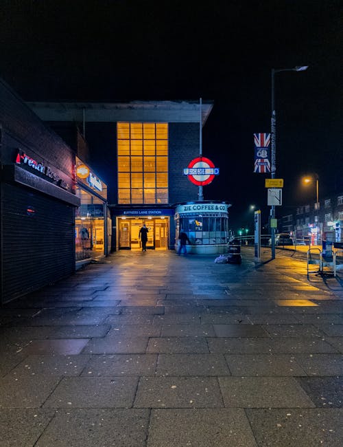 A nighttime view of the exterior of Rayners Lane Station, showing the illuminated entrance with large glass panels and visible signage, including the London Underground roundel and directional signs. The station is located on a paved sidewalk area with some reflections from recent rain. To the right, a lamppost and a flagpole with the Union Jack flag are visible, along with a small kiosk marked 'ZIE Coffee & Co' near the station entrance. The environment appears calm with minimal foot traffic, and nearby buildings are lit, highlighting an urban setting suitable for home relocation and packing activities related to furniture transport and moving logistics, as handled by Man with Van Rayners Lane.