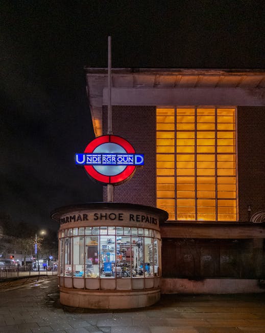 Nighttime exterior scene of a building on Alexandra Avenue in Rayners Lane, showing a small circular shoe repair shop with large glass windows displaying various footwear and tools inside. Above the shop, a partially illuminated sign reads 'UNDERGROUND' in red, white, and blue, indicating proximity to Rayners Lane underground station. The building features a large brick wall with a prominent rectangular window partitioned into smaller panes, emitting warm yellow light. Adjacent to the shop, there is a visible sidewalk and street pavement, with faint streetlights and parked vehicles in the background. This setting reflects an urban area suitable for house removals and furniture transport, with the shop being part of a typical residential and commercial neighborhood, and the scene captured during evening hours, highlighting the residential and local business environment where a professional removals service such as Man with Van Rayners Lane operates.
