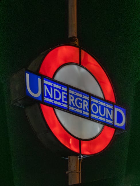 A bright illuminated London Underground roundel sign featuring a red outer ring, a white central circle, and a blue rectangular bar with the word 'UNDERGROUND' in white capital letters. The sign is mounted on a wooden pole against a dark night sky and is prominently visible as part of a house removal or move related to the area around Alexandra Avenue, Rayners Lane, with the sign likely located near the property being relocated. The sign's illumination highlights its vibrant colours, and it serves as a recognizable symbol of London transportation, supporting the context of furniture transport or packing and moving services provided by Man with Van Rayners Lane during a small flat move.