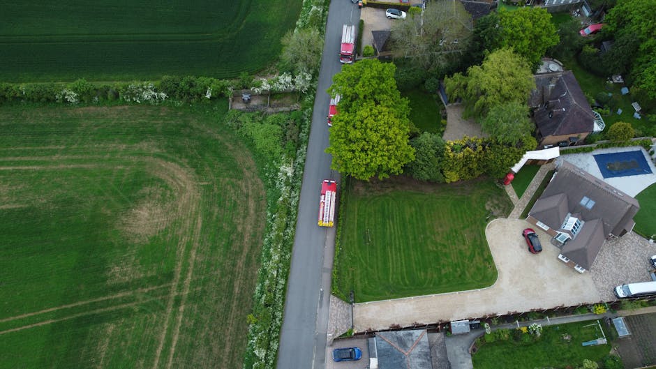 An aerial view of a residential driveway with a red and white moving truck parked on the paved surface, surrounded by green lawns, trees, and residential buildings. Nearby, a garden area with bushes and flowering plants is visible, along with a swimming pool in one backyard. The scene captures the loading or unloading process associated with home relocation services, with the truck positioned for furniture transport or packing and moving activities. The image is taken during daylight, showing clear visibility of the property's exterior environment, contributing to the context of house removals and furniture transport facilitated by Man with Van Rayners Lane.