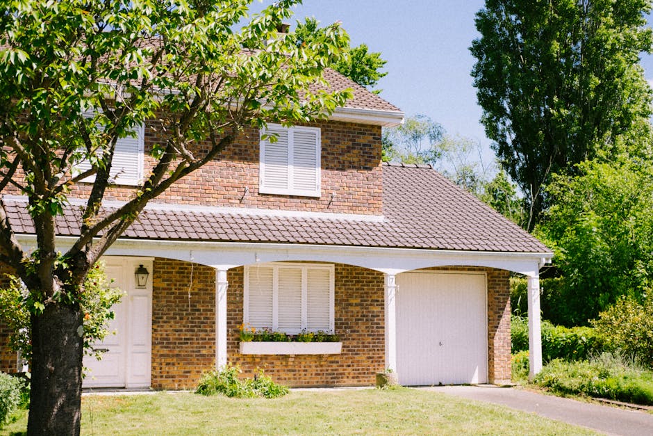A two-story brick house with a pitched tiled roof and white window shutters, situated in a suburban setting with green trees and a well-maintained lawn. The ground floor features a white front door with a lantern-style light beside it, and a large window with closed shutters adorned with a flower box containing small plants. Next to the house, a driveway leads to a single garage door, and a concrete pathway runs along the front. A person from Man with Van Rayners Lane is visible loading a large, flat-packed cardboard box onto a trolley, preparing for house removal or moving process. The scene is illuminated by natural daylight, with bright sky and shadows cast by the trees, indicating a clear, sunny day, and the environment suggests an organized home relocation setup with furniture transport and packing materials ready for safe moving logistics.